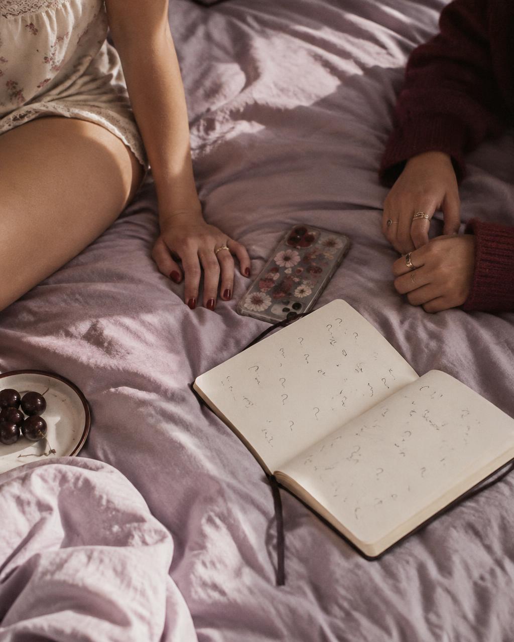 Two young women's hands resting on a lavender bedspread beside a phone and an open notebook filled with handwritten question marks